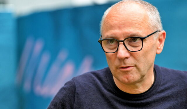 Older man with glasses in a dark sweater, looking to the side, outdoors in front of a blue backdrop.