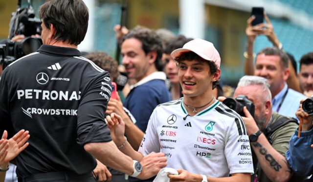 Young racer in a white Petronas shirt smiles while shaking hands with a man in a black team shirt at a crowded media event with photographers around.