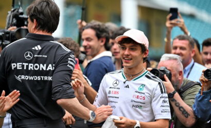 Mercedes-AMG Petronas driver in a white team shirt and pink cap smiles while shaking hands with a team member amid cheering photographers and fans