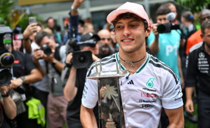 Young male race car driver in a white Petronas shirt and pink cap holds a tall trophy as photographers cluster around him.
