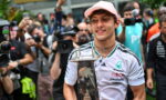 Young man wearing a pink cap and white racing jersey holds a tall trophy while photographers surround him.