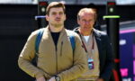 Two men stand outdoors near an event setup; the younger man in a tan jacket with a blue backpack looks ahead, wearing a wristwatch.