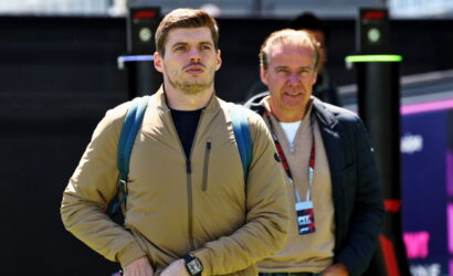 Two men stand outdoors near an event setup; the younger man in a tan jacket with a blue backpack looks ahead, wearing a wristwatch.