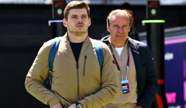 Two men stand outdoors near an event setup; the younger man in a tan jacket with a blue backpack looks ahead, wearing a wristwatch.
