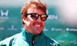 Smiling man wearing dark sunglasses and a teal zip-up jacket at an outdoor event backdrop.