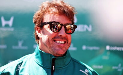 Smiling man wearing dark sunglasses and a teal zip-up jacket at an outdoor event backdrop.