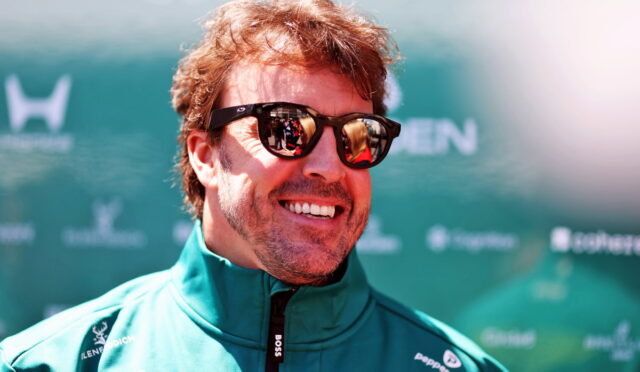Smiling man wearing dark sunglasses and a teal zip-up jacket at an outdoor event backdrop.