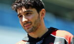 Close-up portrait of a man with dark hair and stubble, outdoors in a red and black sports jacket with sunglasses hanging from his neck.