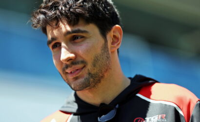 Close-up portrait of a man with dark hair and stubble, outdoors in a red and black sports jacket with sunglasses hanging from his neck.