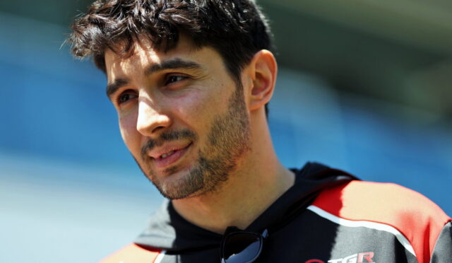 Close-up portrait of a man with dark hair and stubble, outdoors in a red and black sports jacket with sunglasses hanging from his neck.