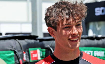 Young man with short wavy brown hair smiling outdoors, wearing a red and black sports jersey at a race event.