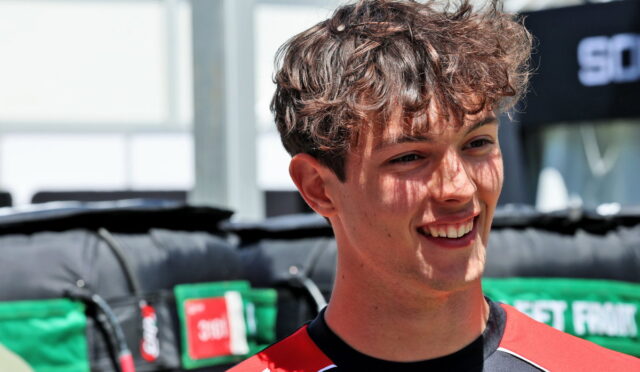Young man with short wavy brown hair smiling outdoors, wearing a red and black sports jersey at a race event.