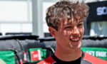 Smiling young man in a red-and-black race jersey at an outdoor event, with padded barriers in the background.