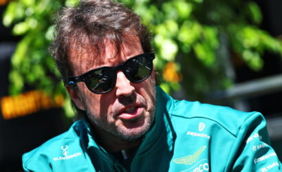 Man wearing a teal race-team jacket and black sunglasses outdoors, looking to the side.
