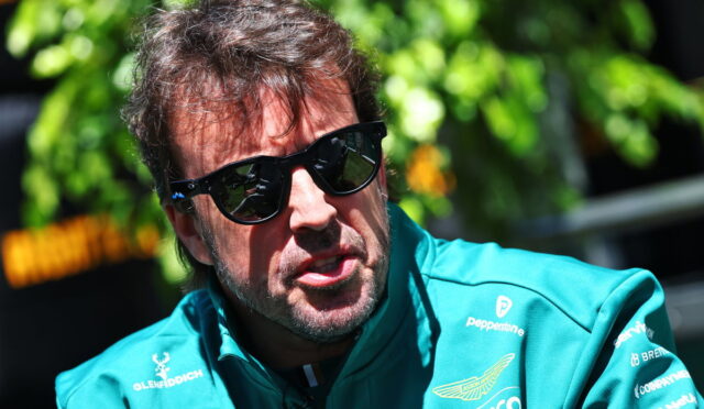Man wearing a teal race-team jacket and black sunglasses outdoors, looking to the side.