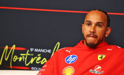 Portrait of a man in a red Ferrari jacket with sponsor logos (HP, Shell) on a dark backdrop, looking thoughtful.