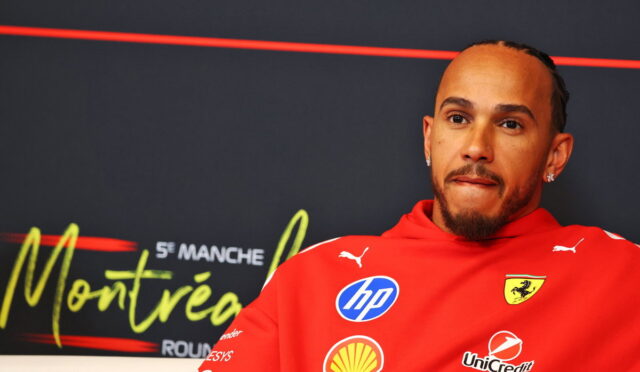 Portrait of a man in a red Ferrari jacket with sponsor logos (HP, Shell) on a dark backdrop, looking thoughtful.