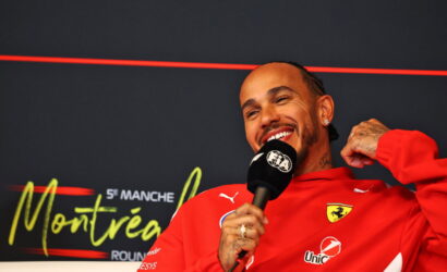 Smiling man in a red Ferrari jacket being interviewed, holding a microphone in front of a dark backdrop.
