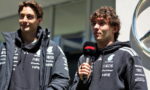 Two men in dark team jackets being interviewed; one holds a microphone with a red logo, near a car dealership backdrop.
