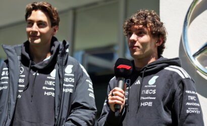 Two men in dark team jackets being interviewed; one holds a microphone with a red logo, near a car dealership backdrop.