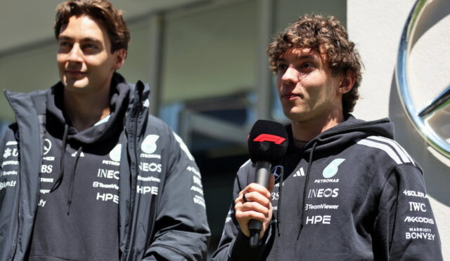 Two men in dark team jackets being interviewed; one holds a microphone with a red logo, near a car dealership backdrop.