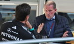 Two men sit at a table in a conversation; one wears a Mercedes Petronas racing jacket, the other gestures while talking.