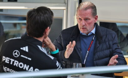 Two men sit at a table in a conversation; one wears a Mercedes Petronas racing jacket, the other gestures while talking.