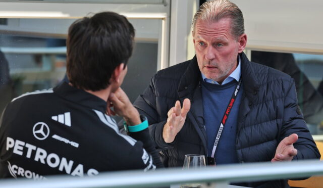 Two men sit at a table in a conversation; one wears a Mercedes Petronas racing jacket, the other gestures while talking.
