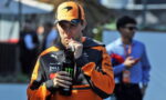 Racer in orange and black gear drinks from a black bottle with a Monster logo outdoors, cap on orange helmet nearby.