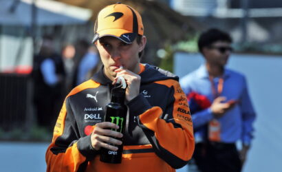 Racer in orange and black gear drinks from a black bottle with a Monster logo outdoors, cap on orange helmet nearby.