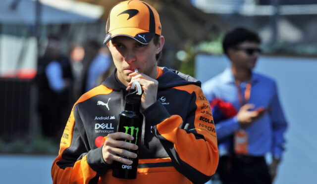 Racer in orange and black gear drinks from a black bottle with a Monster logo outdoors, cap on orange helmet nearby.