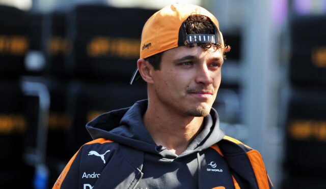 Young man in an orange cap and dark team jacket, looking to the right at a racing event.