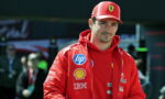 Male race car driver in a red Ferrari jacket and cap, sponsor logos visible, standing in the pit area.