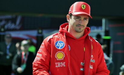Male race car driver in a red Ferrari jacket and cap, sponsor logos visible, standing in the pit area.