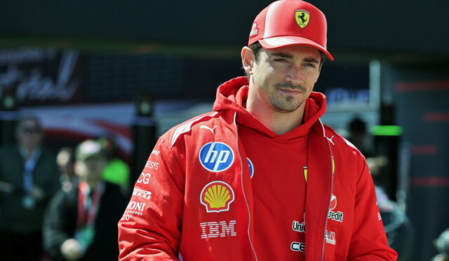 Male race car driver in a red Ferrari jacket and cap, sponsor logos visible, standing in the pit area.