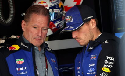 Two Oracle Red Bull Racing team members in blue jackets discuss something in the garage, one wearing a cap with sponsor logos