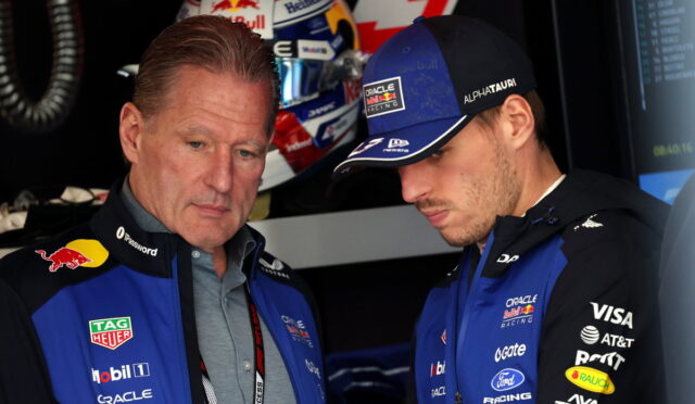 Two Oracle Red Bull Racing team members in blue jackets discuss something in the garage, one wearing a cap with sponsor logos