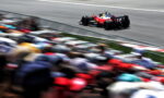 Red formula-style race car speeding along a track with a colorful, blurred crowd in the foreground, emphasizing speed.