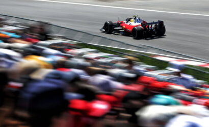 Red formula-style race car speeding along a track with a colorful, blurred crowd in the foreground, emphasizing speed.