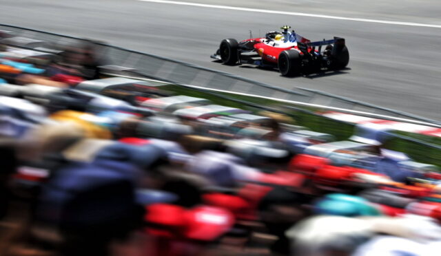 Red formula-style race car speeding along a track with a colorful, blurred crowd in the foreground, emphasizing speed.