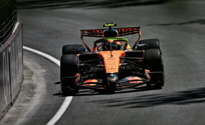 Orange Formula 1 car rounding a left-hand curve on a race track, barrier to the left, driver wearing a bright helmet. Silhouette and sponsor logos visible on the car's bodywork.