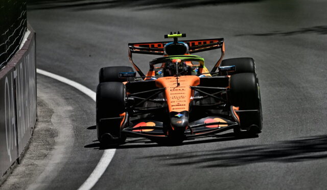 Orange Formula 1 car rounding a left-hand curve on a race track, barrier to the left, driver wearing a bright helmet. Silhouette and sponsor logos visible on the car's bodywork.