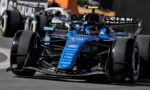 Blue Formula 1 car taking a tight corner, sponsor logos visible on the nose and wings, racing closely with another car behind it.