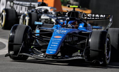 Blue Formula 1 car taking a tight corner, sponsor logos visible on the nose and wings, racing closely with another car behind it.