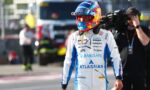 Race car driver in a white-and-blue suit with Barclays and Atlassian logos, wearing a colorful helmet, walking in the pit lane with crew in the background.