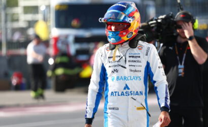 Race car driver in a white-and-blue suit with Barclays and Atlassian logos, wearing a colorful helmet, walking in the pit lane with crew in the background.