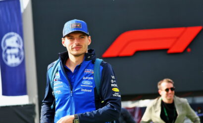 Male Formula 1 team member in blue jacket and cap, carrying a backpack, standing outdoors near a large red F1 logo.