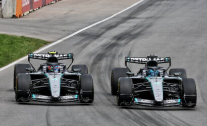 Two Mercedes Formula One race cars side by side on a track during a race, tires smoking slightly at the corner of a circuit road edge, with barriers on the side.
