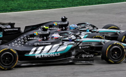 Three black Mercedes Formula 1 race cars with Petronas livery speeding on a track, sponsors visible on the chassis and helmets.