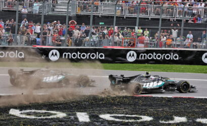 Two Formula 1 cars race off the track as dust and debris kick up near the barrier, with a packed grandstand and Motorola banners in the background.